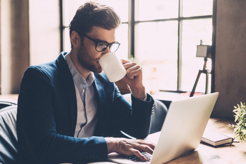Enjoying fresh coffee at work. Confident young man in smart casual wear working on laptop and drinking some hot drink while sitting at his working place in office