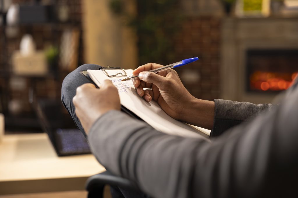 Black male psychologist writing in notepad, taking notes at private clinic. African american therapist with pen in hand, documenting on clipboard during session in brick wall office.
