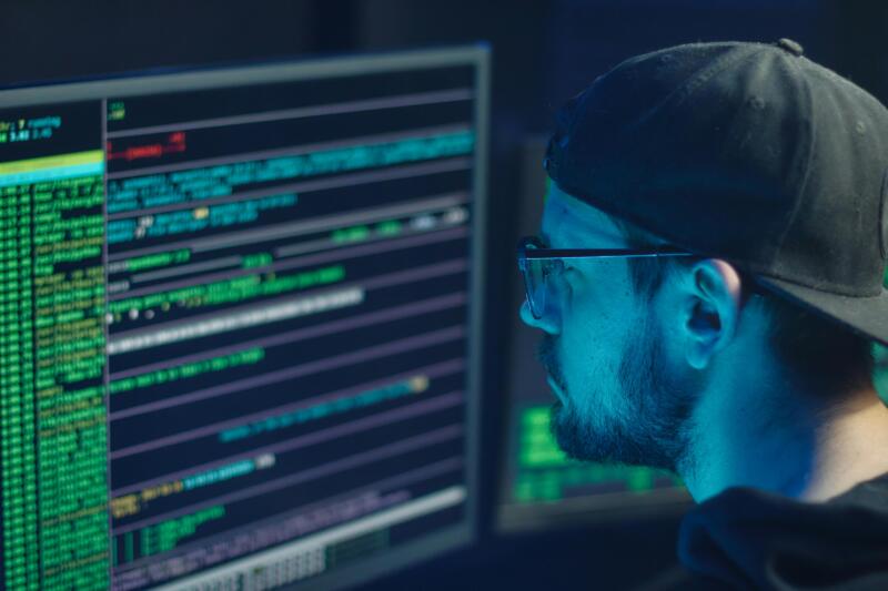 Man wearing a cap and glasses analyzing lines of code on multiple computer monitors in a dimly lit room, symbolizing cybersecurity and data risk management